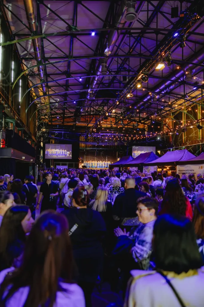 Blick über die Gästemenge in einer Industriehalle mit Bühnen-Livestream auf Leinwänden und Food-Ständen bei einem Corporate Event.