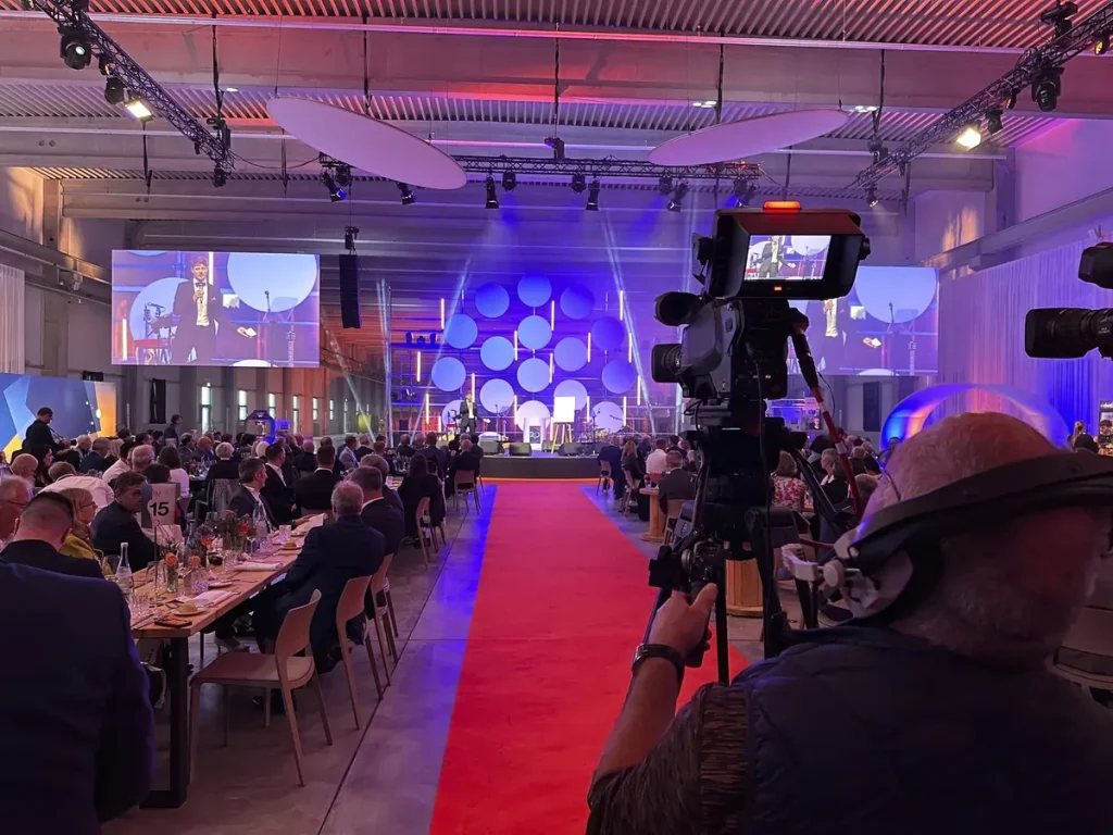 Gästetafel mit rotem Teppich und Bühnenprogramm auf Leinwänden, im Vordergrund ein Kamerateam bei der Liveübertragung der VEROPE-Jubiläumsfeier.
