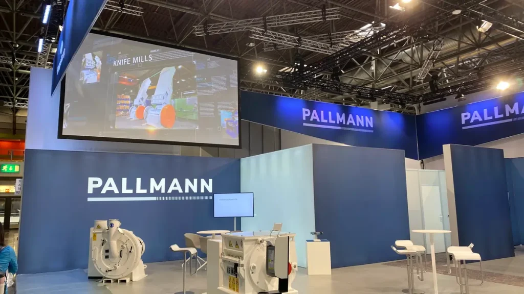 Pallmann-Messestand mit Maschinenexponaten und Präsentationsleinwand in einer Ausstellungshalle.