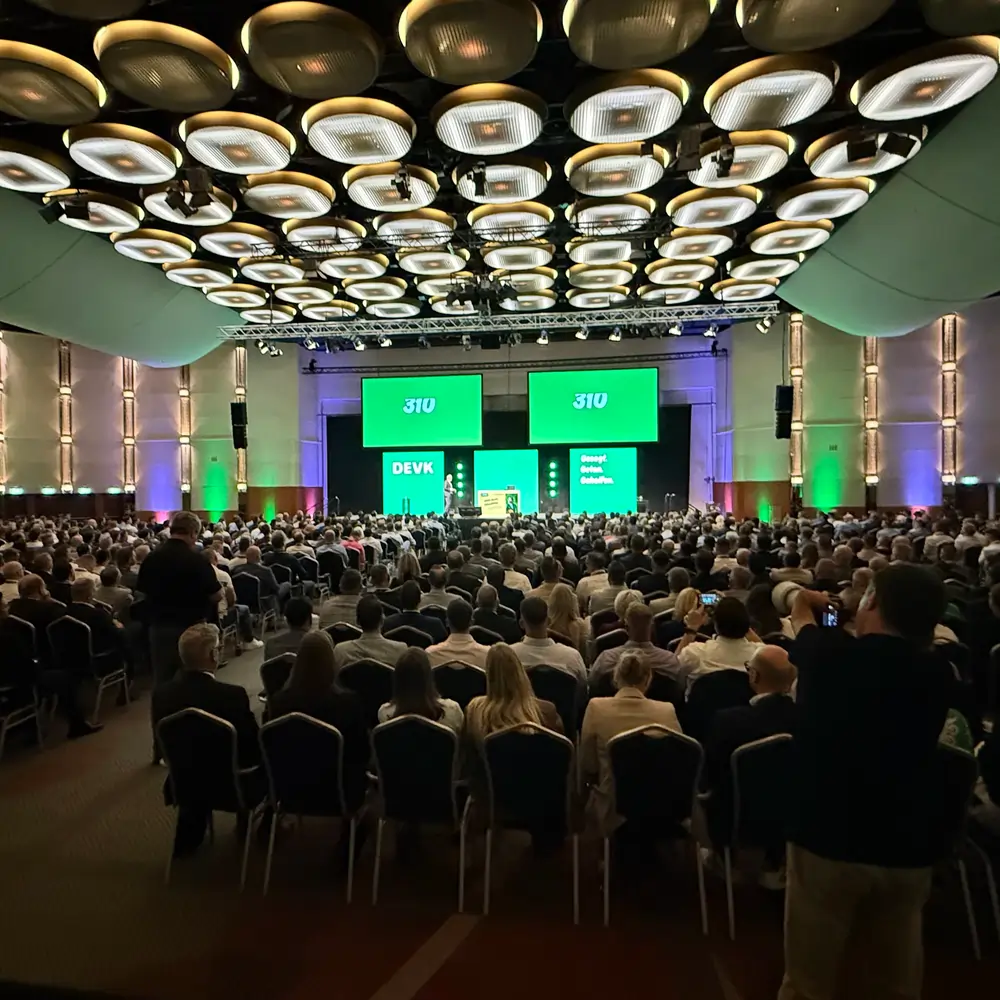 Vollbesetzter Konferenzsaal mit DEVK-Bühnenprogramm auf Videowänden.